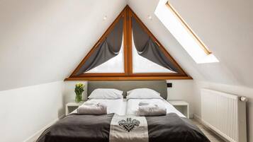 A bedroom with a sloped ceiling, a bed with a dark headboard, a bedside table with a vase of flowers, and a window with curtains.