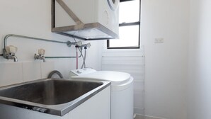 A kitchen with a stainless steel sink, a white washing machine, and a window with blinds.