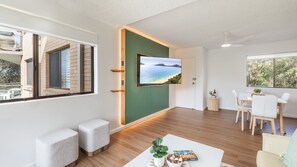 A modern living room with a large TV mounted on a green wall, a dining area with white furniture, and a view of a beach through a window.