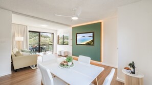 A modern living room with a dining area, a ceiling fan, and a large window with a view of greenery.