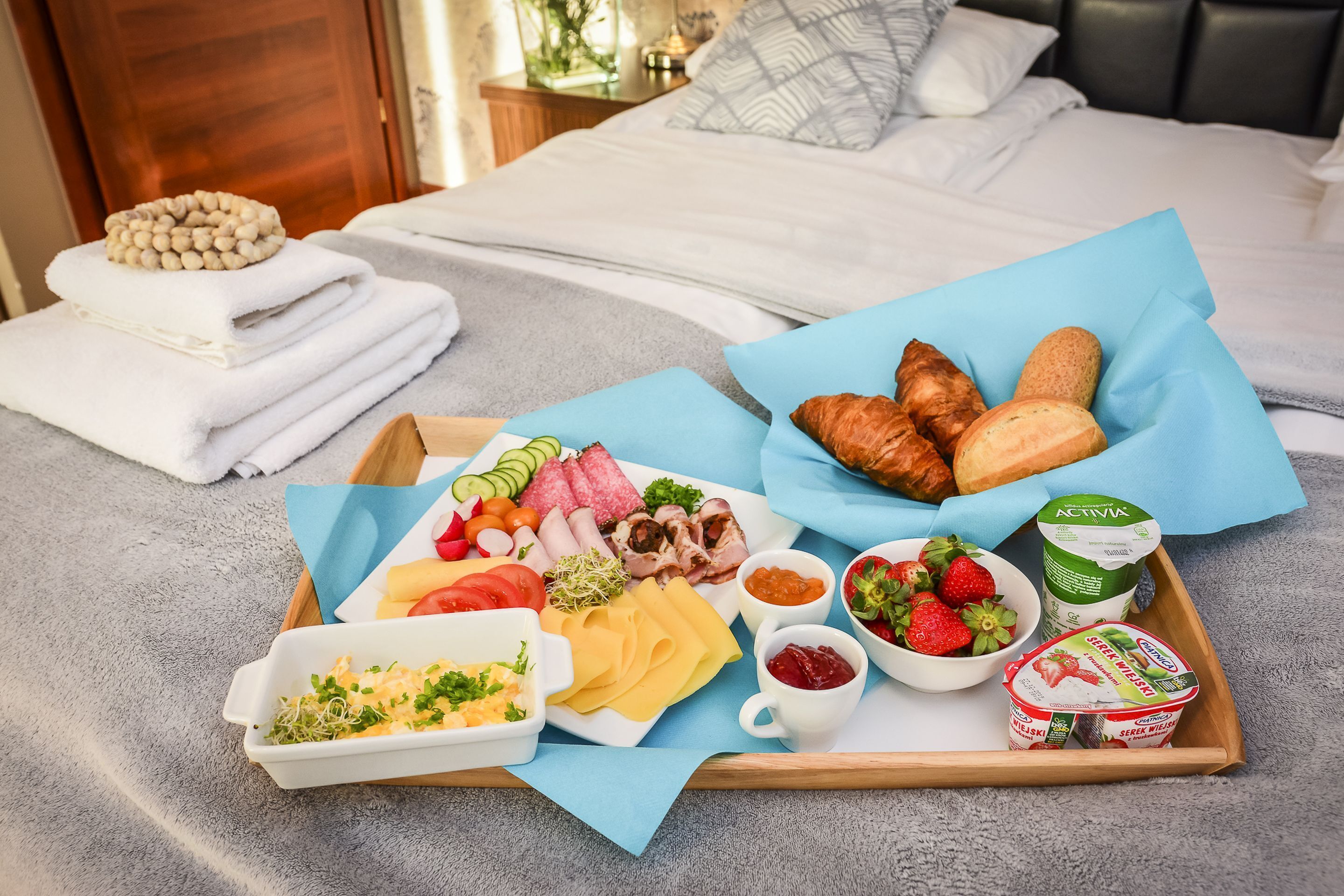 Un plateau de petit-déjeuner contenant divers aliments, notamment du pain, du fromage, de la charcuterie et du yaourt, posé sur un lit recouvert d’une couverture bleue.