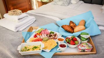 Un plateau de petit-déjeuner contenant divers aliments, notamment du pain, du fromage, de la charcuterie et du yaourt, posé sur un lit recouvert d’une couverture bleue.