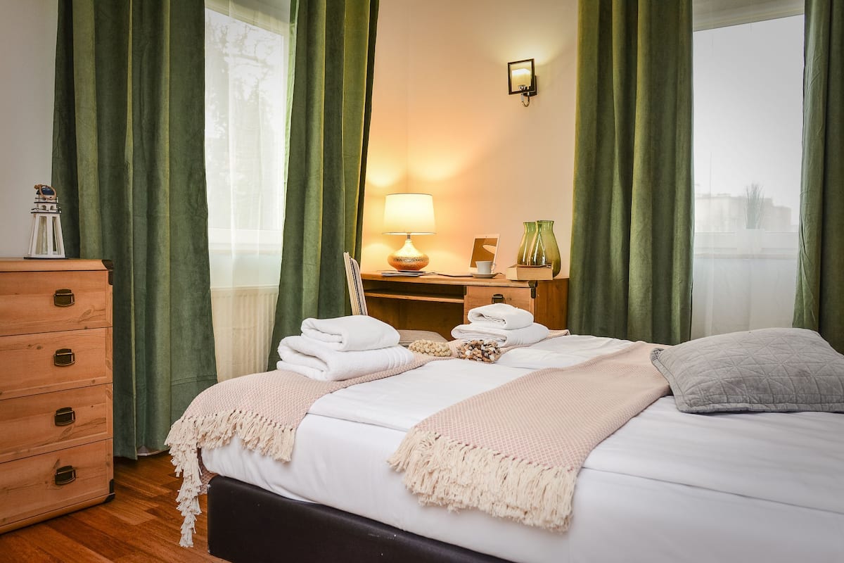A neatly made bed with white linens and a beige throw blanket, a wooden dresser, a bedside table with a lamp, and a view of a cityscape through a window with green curtains.