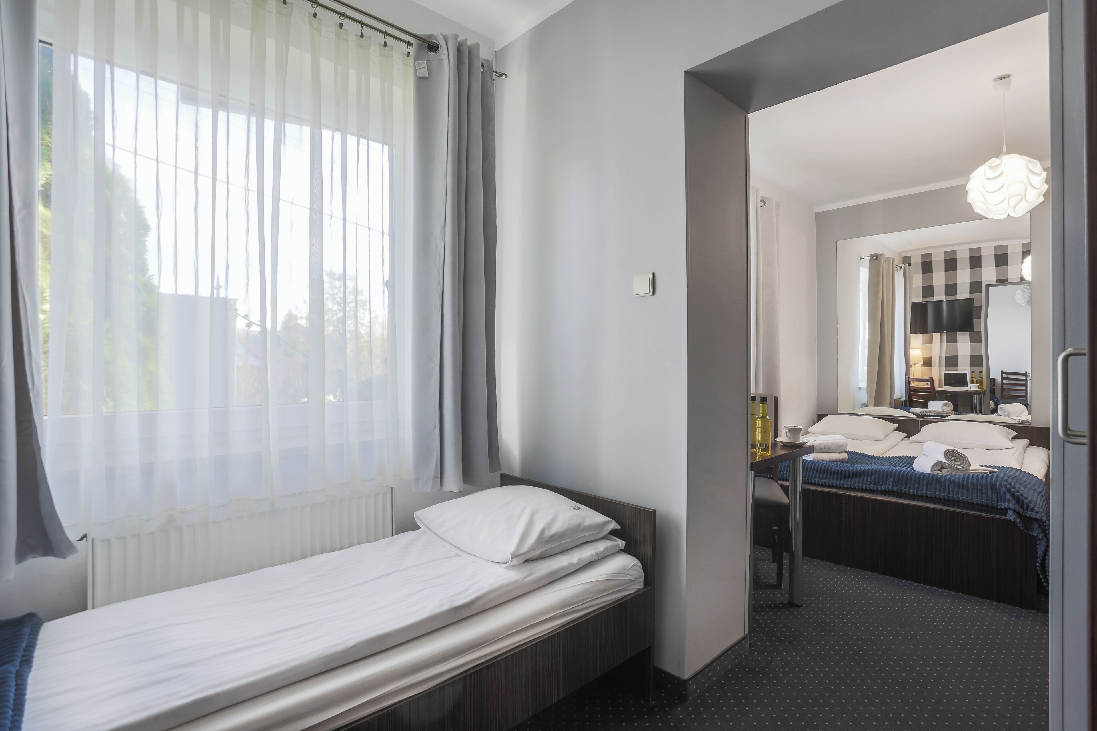 A hotel room with a single bed, a bedside table with a lamp, and a view of another room through an open door.