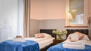 A hotel room with two beds, each with a blue bedspread, white linens, and decorative pillows. There is a nightstand with a lamp and a vase with flowers. - Willa Bryza (Gdynia)