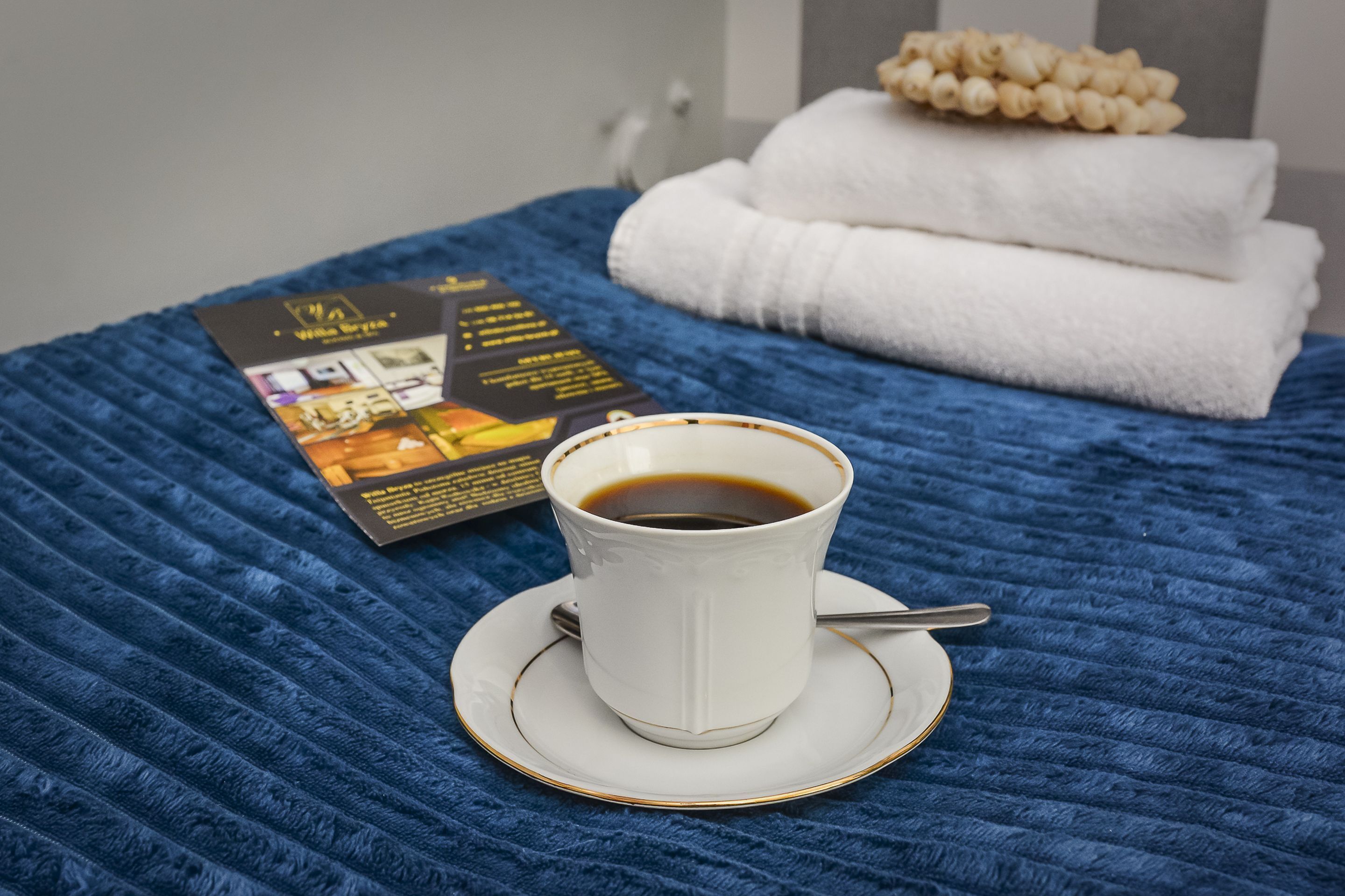 Uma chávena de café branca sobre um pires, um folheto e toalhas dobradas sobre um cobreleiro azul texturizado.