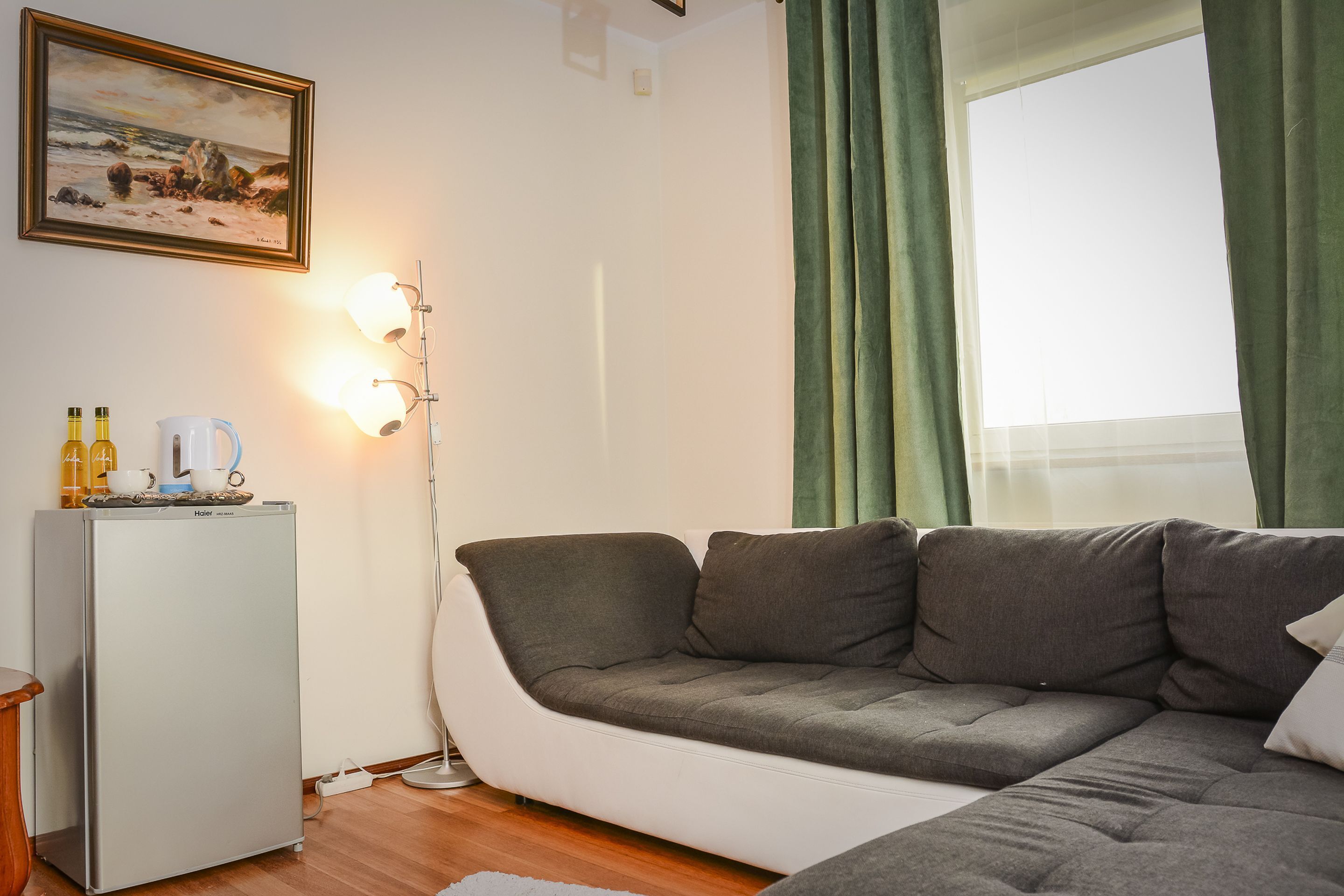 A living room with a grey sofa, a small white fridge, and a painting on the wall.