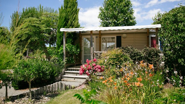 A small wooden cabin with a porch, surrounded by a garden with various plants and flowers. - Camping Saint Michel (Courtils)