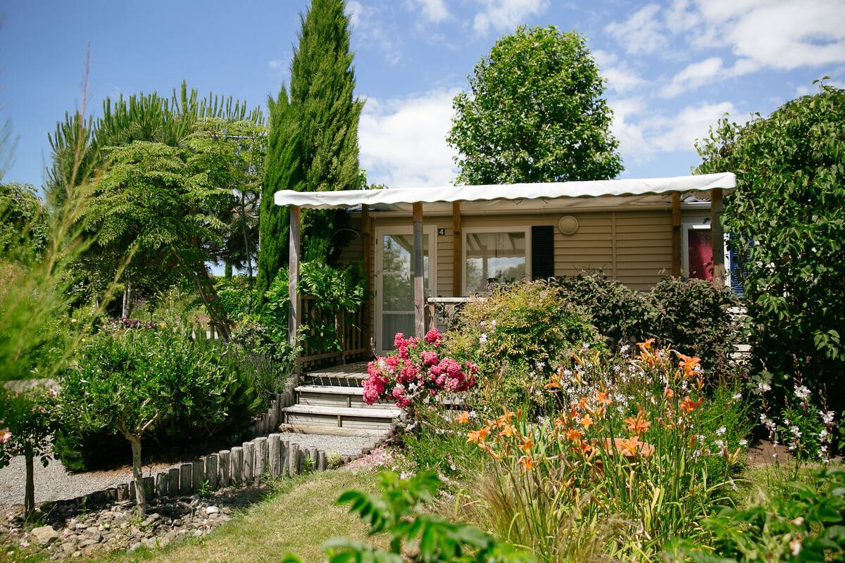 Une petite cabane en bois dotée d’un porche, entourée d’un jardin où poussent différentes plantes et fleurs.