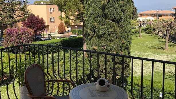 A balcony with a chair and table, a tall tree, and buildings in the background. - Relais Sweet Days (Peschiera del Garda)