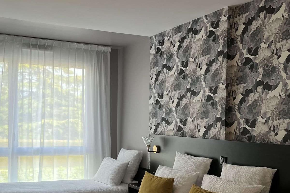 A hotel room with a large bed, a headboard with a floral wallpaper, and a window with sheer curtains.