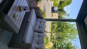 Un sofá de mimbre con cojines, una mesita y una silla de mimbre en un patio con vistas a la entrada de un hotel y vegetación.