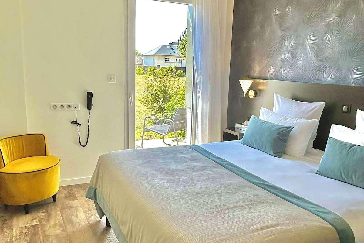 A bedroom with a large bed, a yellow armchair, a bedside table with a lamp, and a view of the outdoors.