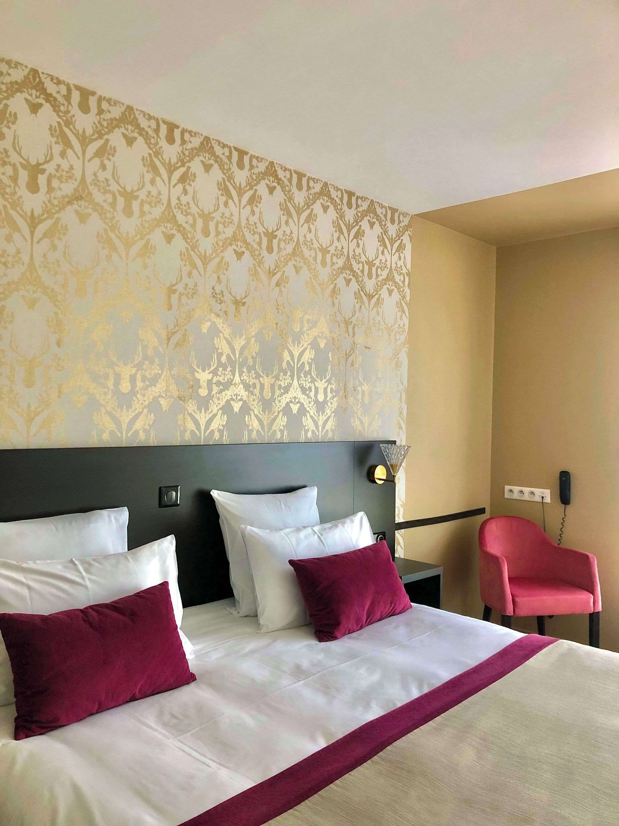 Habitación de hotel con una cama grande, cabecera, mesita de noche, lámpara, teléfono y una silla roja.