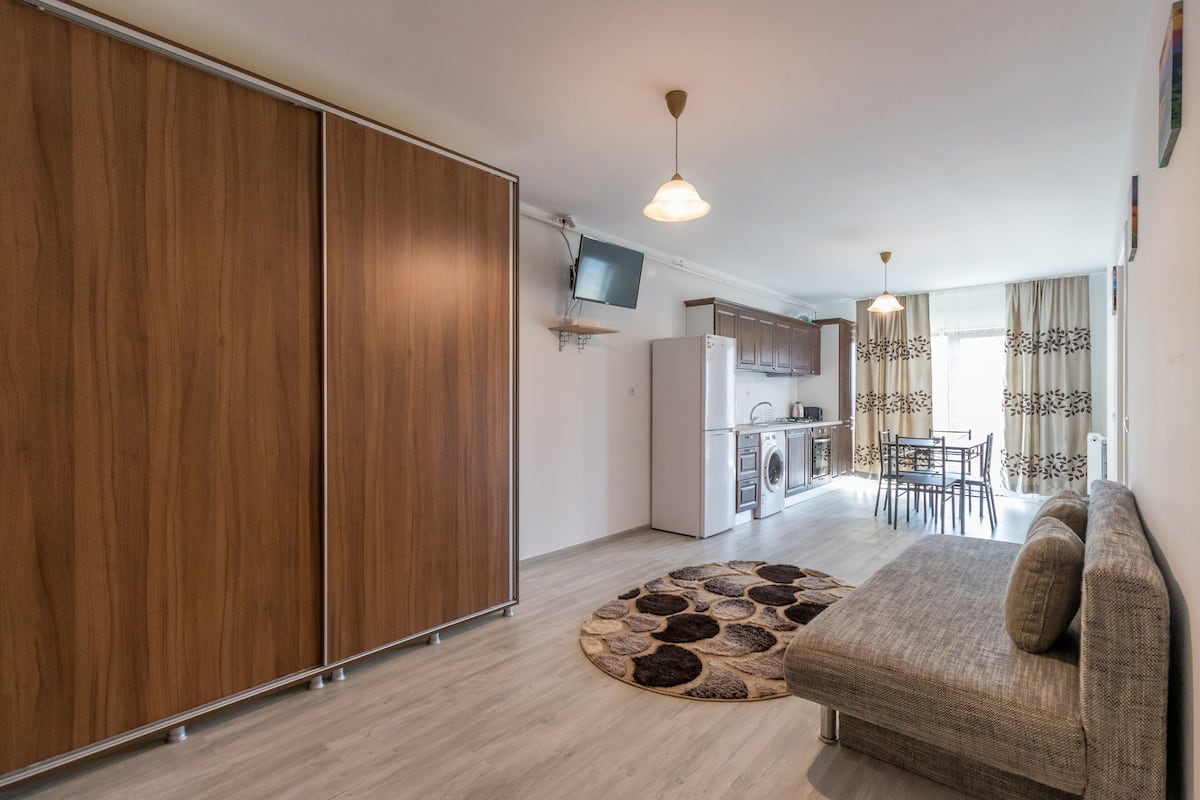 Un apartamento moderno con un armario de madera, una cocina equipada con refrigerador, una zona de comedor con mesa y sillas, y una sala de estar con sofá.