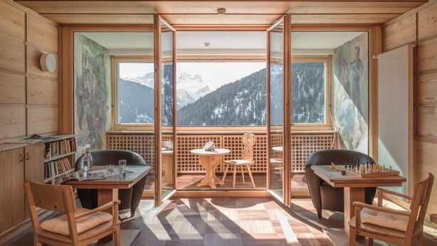 A wooden cabin with a large window overlooking a mountainous landscape. Inside, there is a wooden table with chairs, a bookshelf, and a chessboard. - Hotel Restaurant Capricorns (Muntogna da Schons)