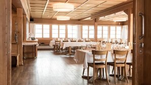 A spacious dining area with wooden flooring and tables set for dining, large windows allowing natural light, and a wooden ceiling with a grid pattern. - Hotel Restaurant Capricorns (Muntogna da Schons)