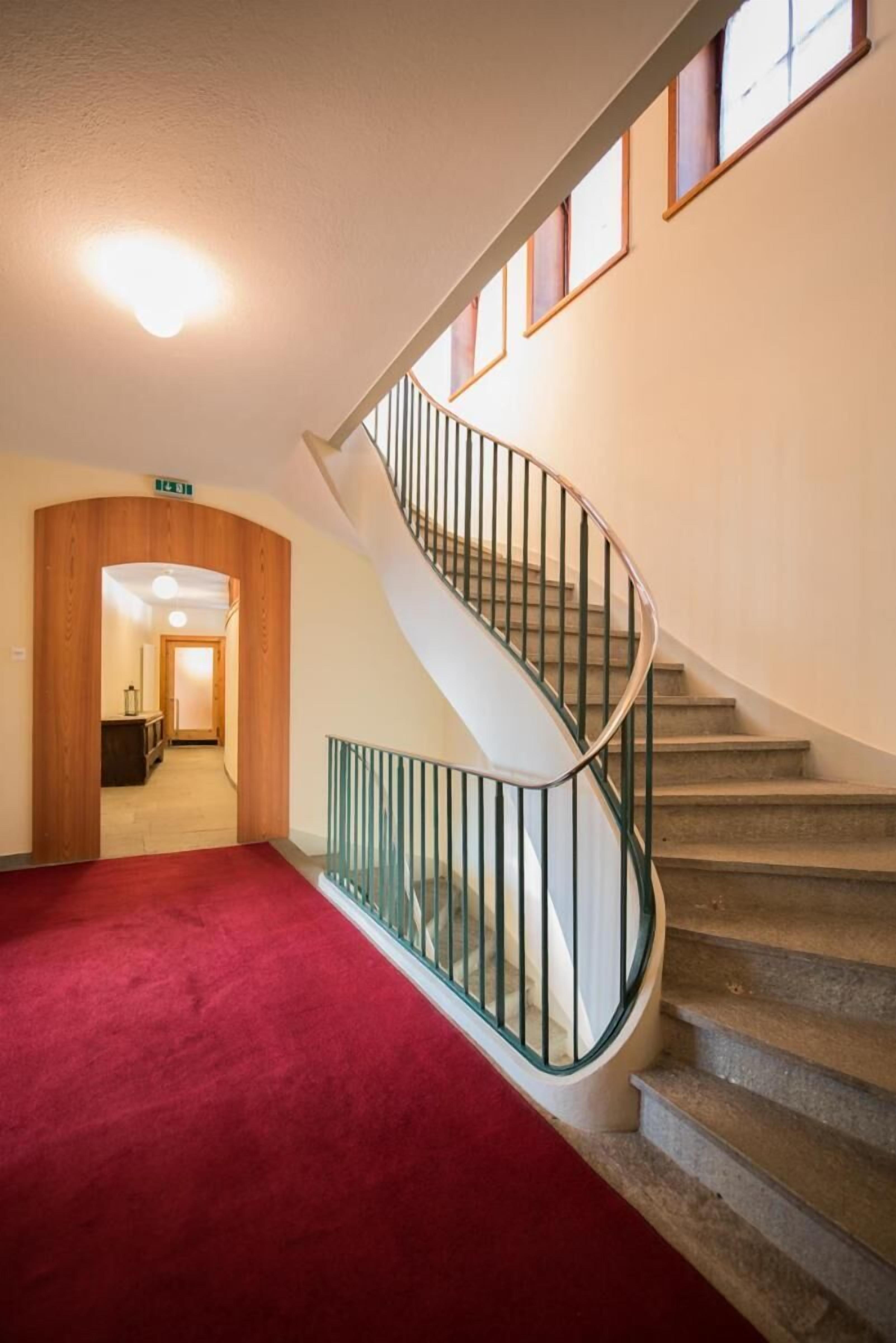 Un escalier avec une rampe en verre, un sol recouvert de moquette et une porte en bois donnant sur une autre pièce.