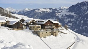 A snow-covered mountain village with traditional wooden buildings and a stone structure, surrounded by snow-covered mountains. - Hotel Restaurant Capricorns (Muntogna da Schons)