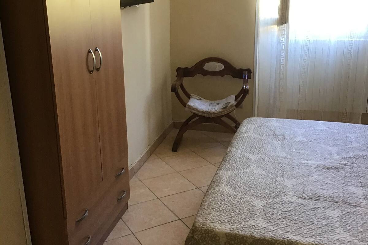 A bedroom with a wooden wardrobe, a chair, a bed with a patterned bedspread, a wall-mounted TV, and a painting.