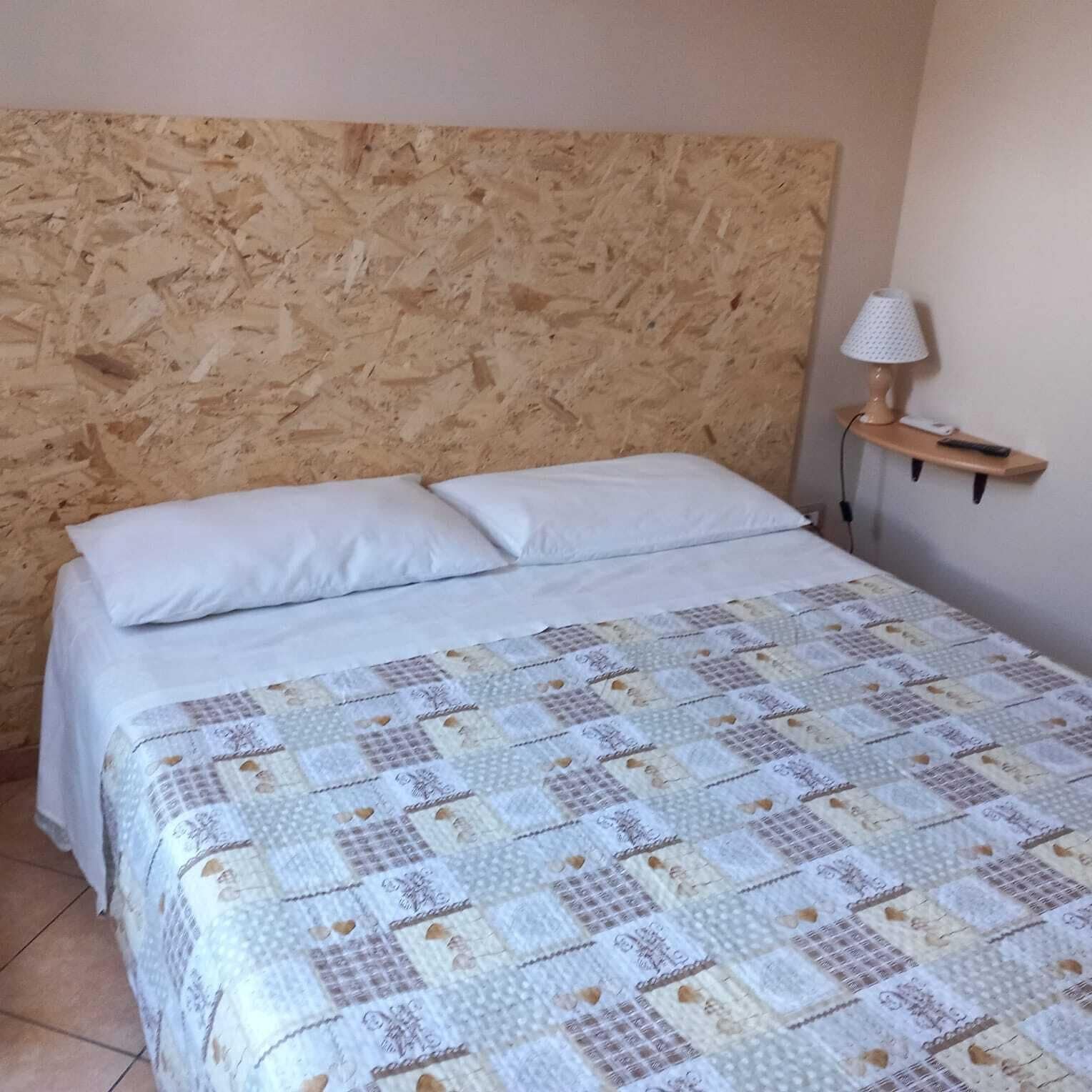 Een slaapkamer met een houten hoofdbord, een gewatteerde spreidek, een bedlampje en een klein plankje met een afstandsbediening.