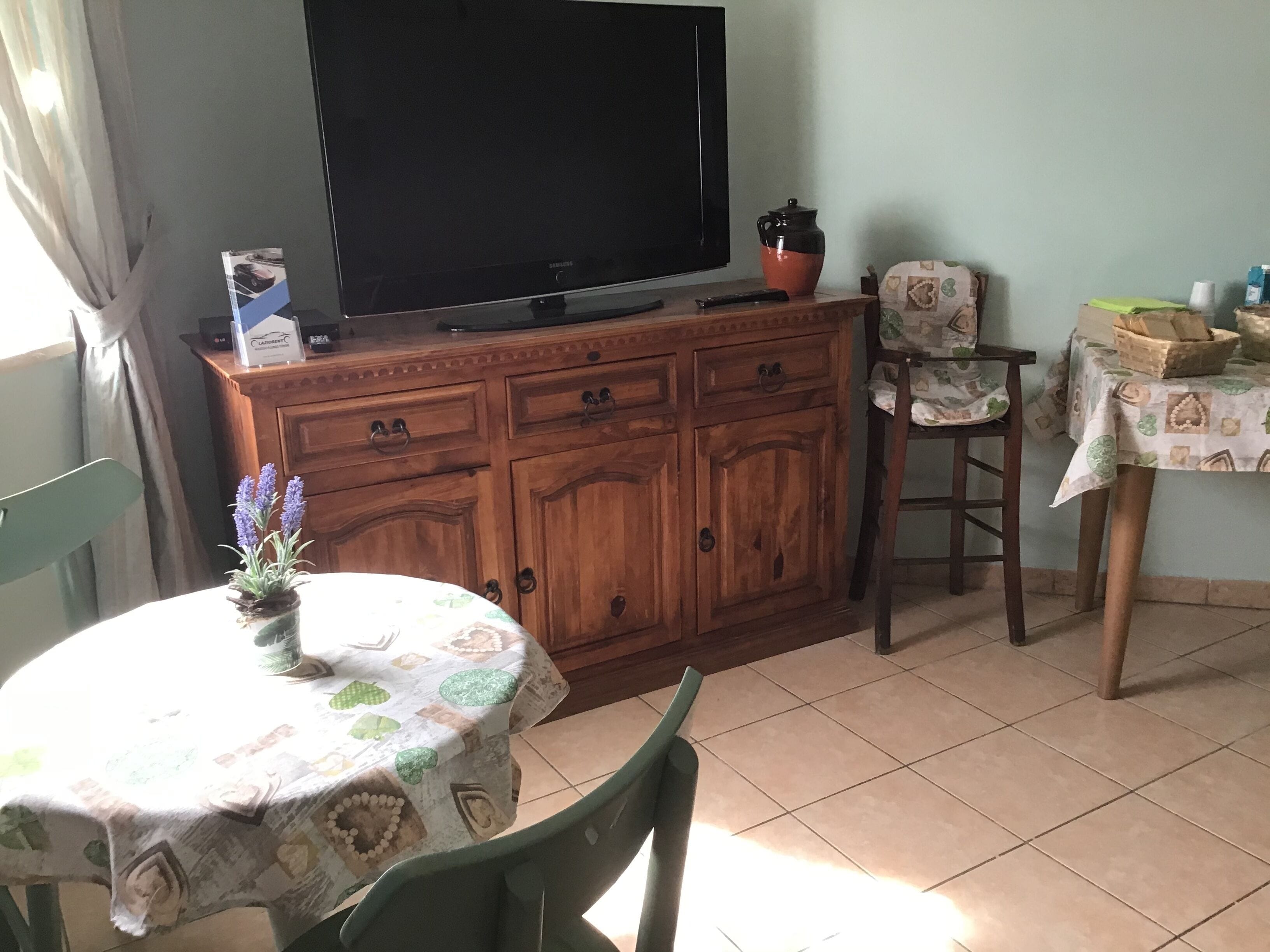 Una habitación con una televisión sobre un mueble de madera, una mesa con mantel estampado, una silla y una mesita con una cesta y una taza.