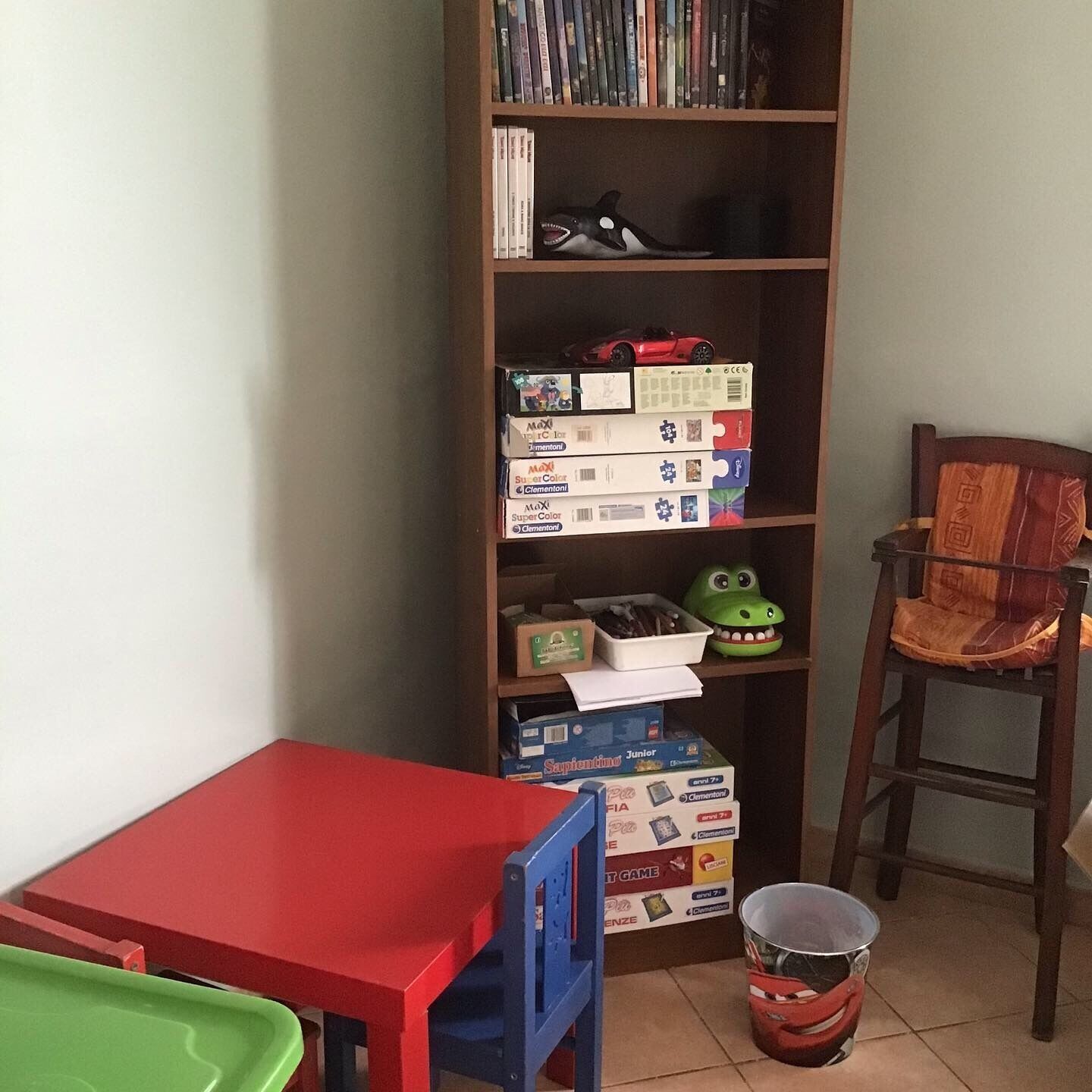 A room with a bookshelf filled with books and toys, a red table, a green chair, and a bucket on the floor.