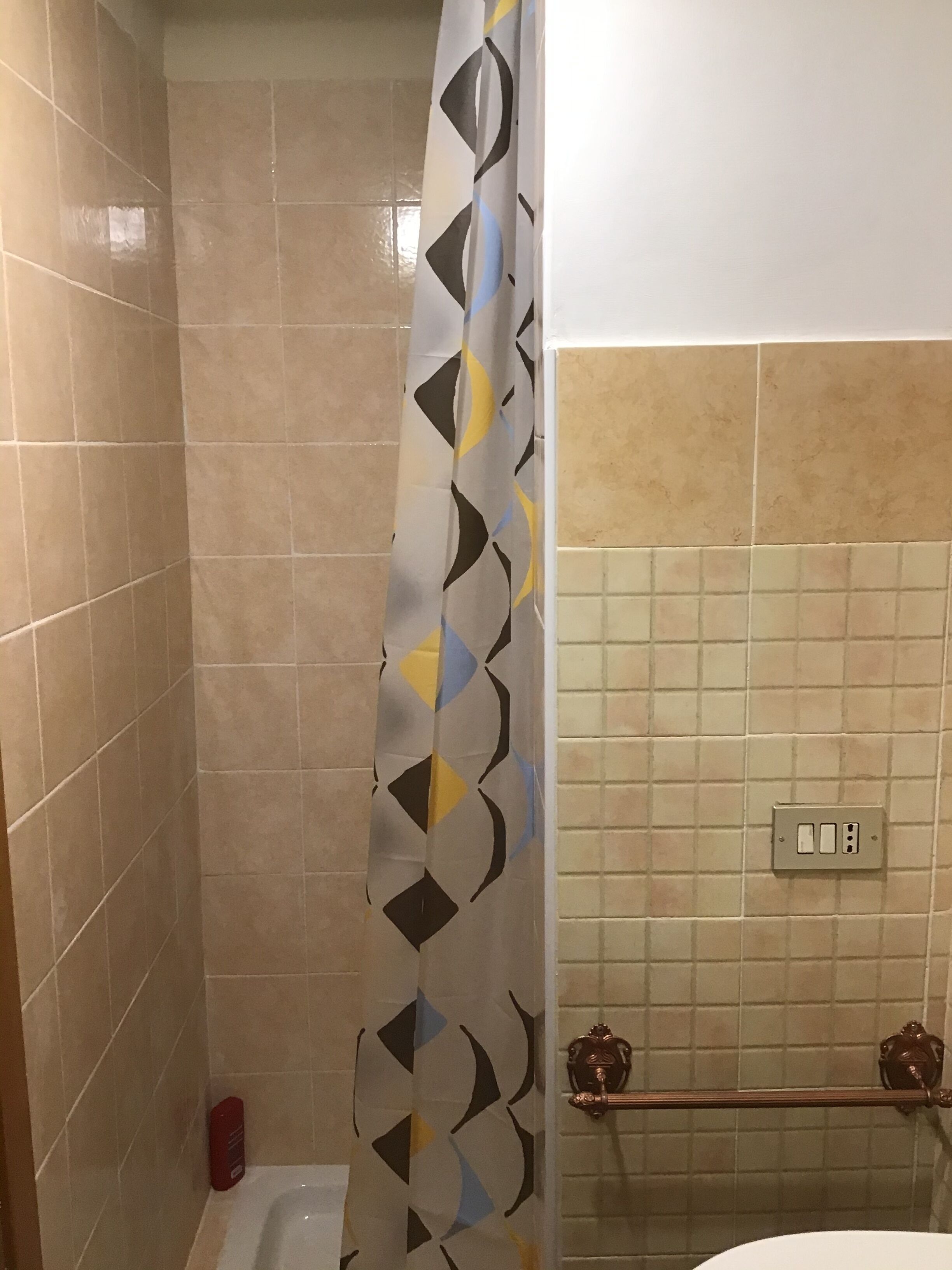 Een badkamer met beige betegelde muren, een douchegordijn met een geometrisch patroon, een toilet, een handdoekhouder en een lichtschakelaar op de muur.