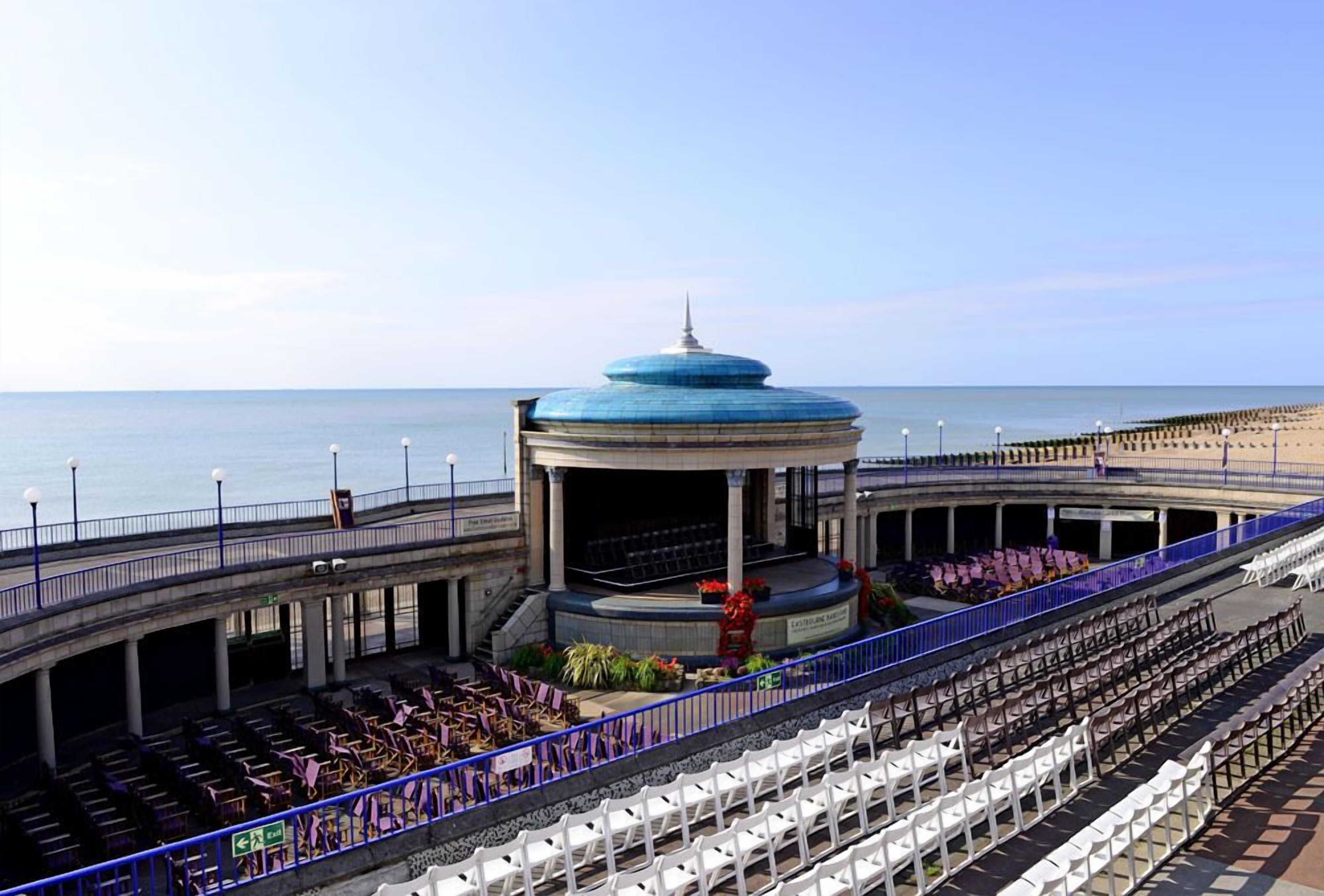 An outdoor amphitheater with tiered seating and a central stage, overlooking a beach.