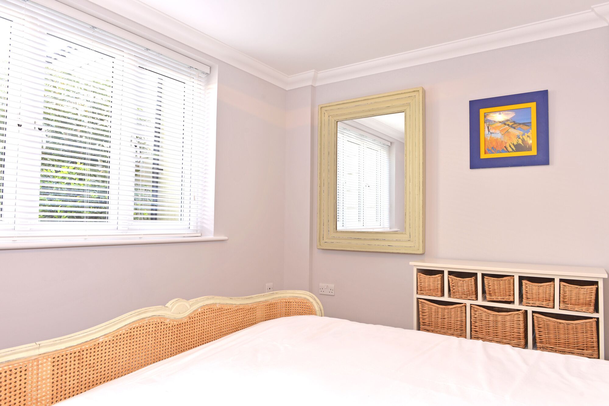 Quarto com cama, janela com persianas, espelho e unidade de armazenamento em cesta fixada na parede.