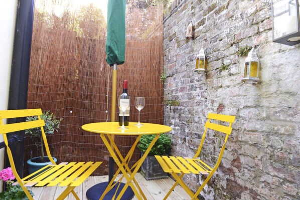 A small outdoor seating area with a round yellow table and two yellow chairs, a bottle of wine, and two glasses on the table.