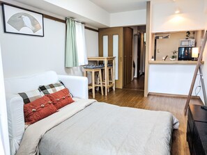 A bedroom with a bed, a chair, a small table, a kitchenette, and a door leading to another room.