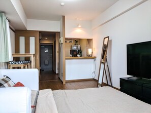 A hotel room with a bed, a desk, a mirror, a TV, and a kitchenette.
