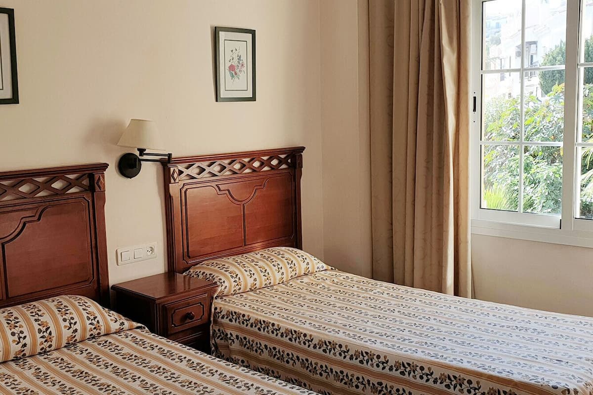 A bedroom with two beds, a nightstand, a wall-mounted lamp, and a window with curtains.