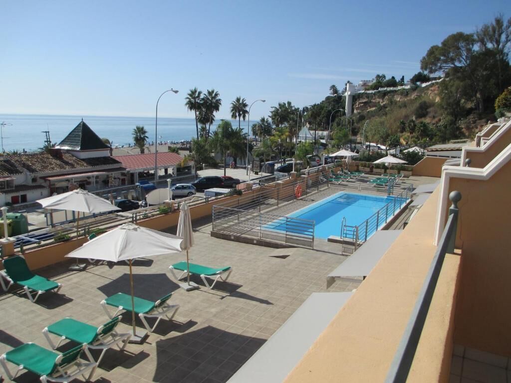 A rooftop pool area with lounge chairs and umbrellas, overlooking a coastal landscape with buildings and a clear sky.