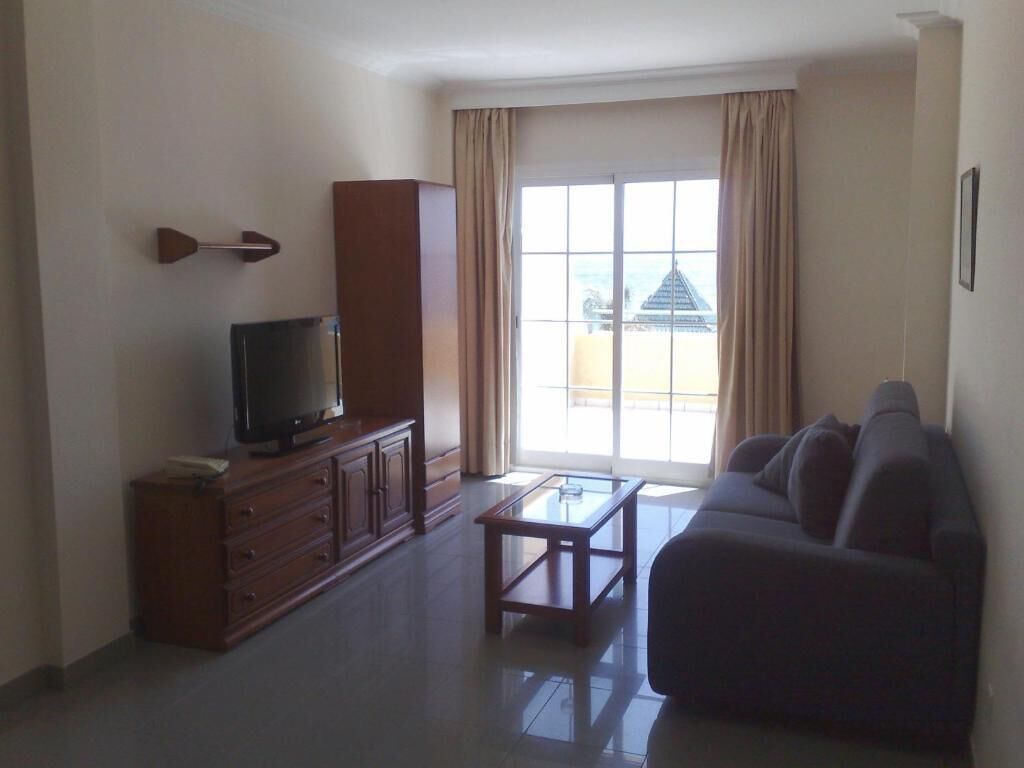 Una sala de estar con televisión sobre un mueble de madera, un sofá, una mesa de centro de vidrio y un ventanal con cortinas.