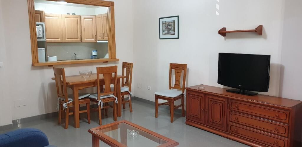 A living room with a wooden dining table, chairs, a glass coffee table, a TV on a wooden stand, and a kitchen area with wooden cabinets and a mirror.