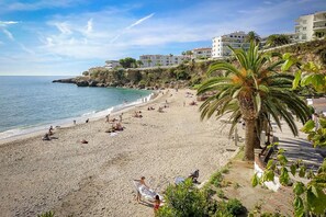 A beach with people sunbathing and swimming, a palm tree, and buildings in the background. - Nerja Bahía Apartamentos (Nerja)