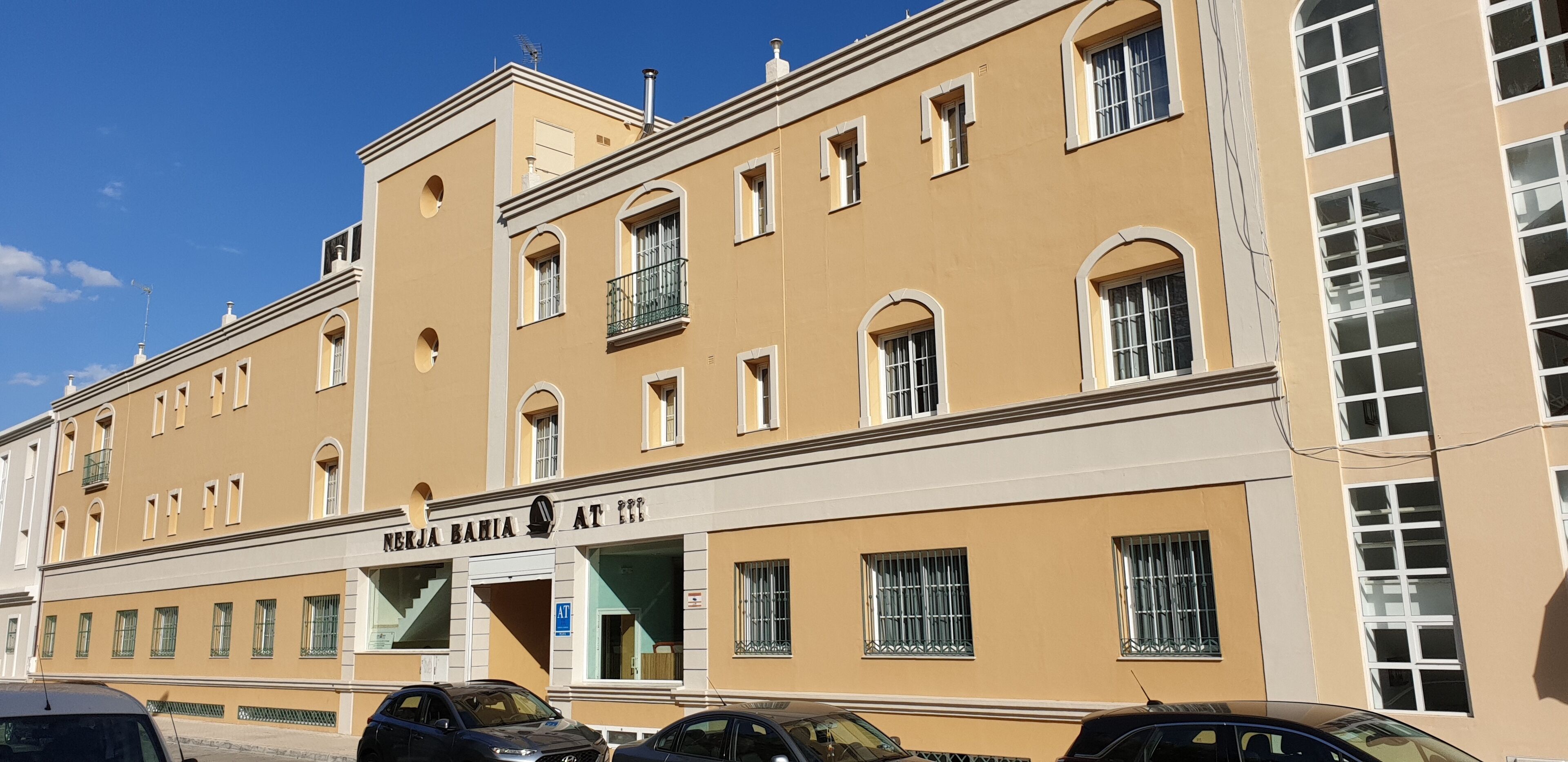 A building with a beige facade, multiple windows, and a sign reading "NERJA BAHIA AT 3".