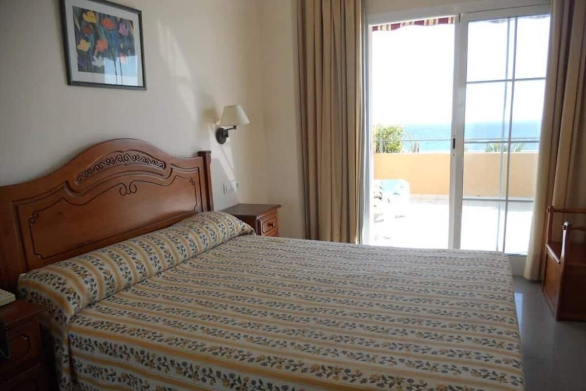 A hotel room with a large bed, a nightstand, a lamp, and a view of the sea through a sliding door.