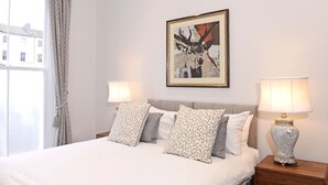 Una cama bien hecha con almohadas estampadas, un cuadro abstracto enmarcado en la pared y dos lámparas de noche sobre mesitas de madera.
