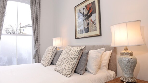 Una cama bien tendida con almohadas estampadas, una obra de arte enmarcada encima y dos lámparas de noche en una mesita.