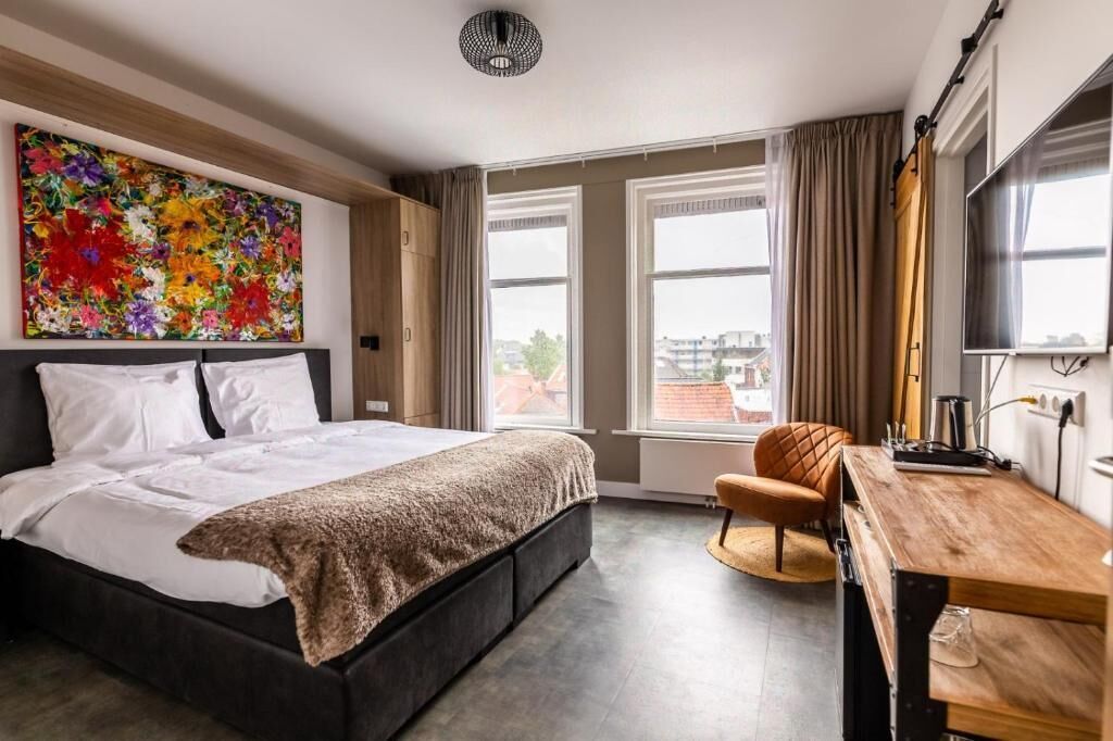 A modern hotel room with a large bed, a desk, a chair, and a large window with curtains.