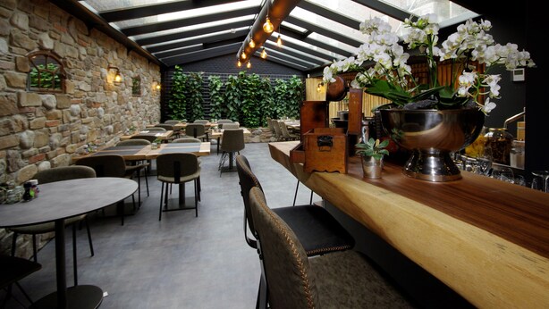 A restaurant with a stone wall, wooden tables, and chairs, and a bar area with a large metal bowl and flowers. - Hotel Restaurant Inn Naeldwyk (Naaldwijk)