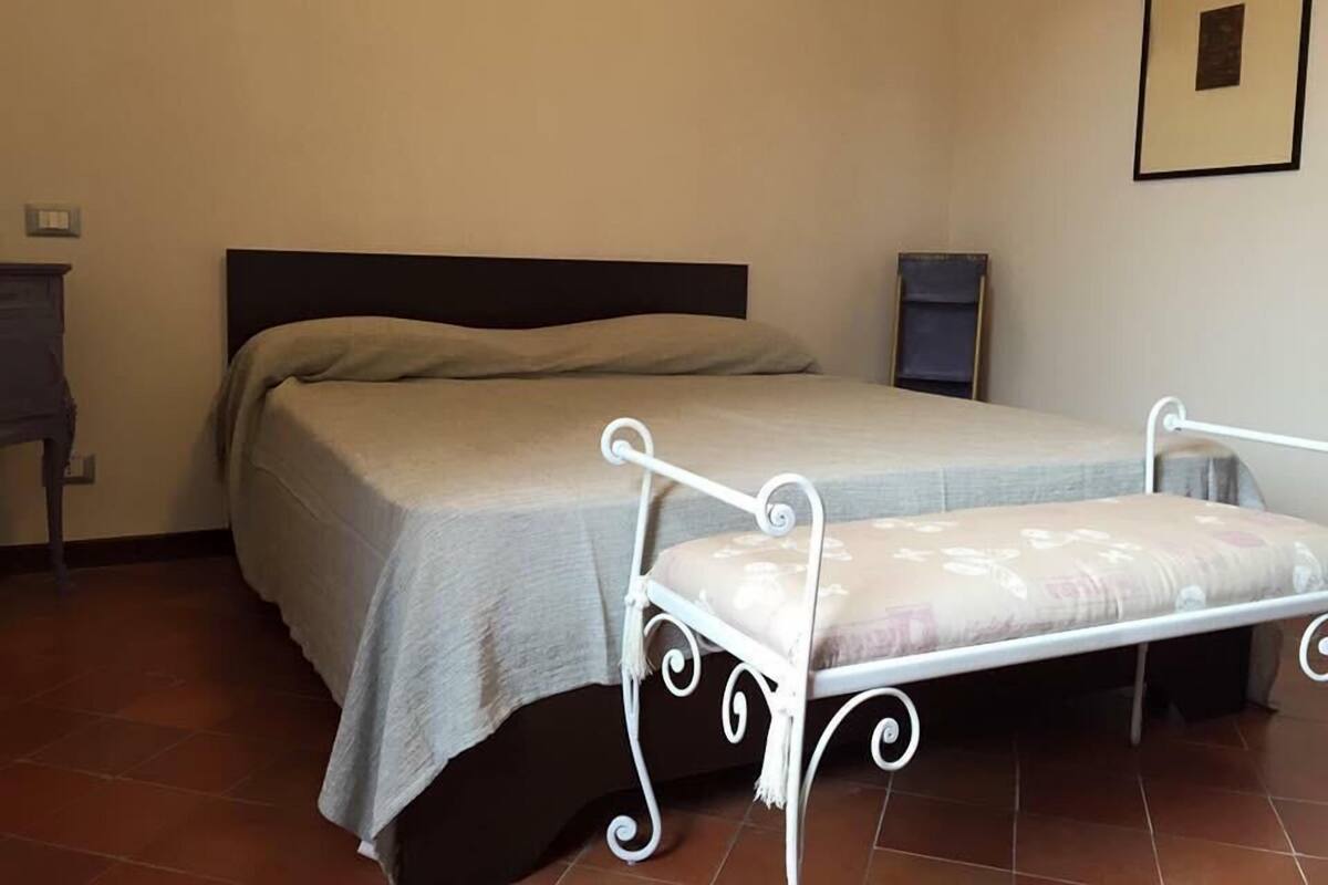 A single bed with a white metal frame and a grey bedspread in a room with tiled flooring and a plain wall.