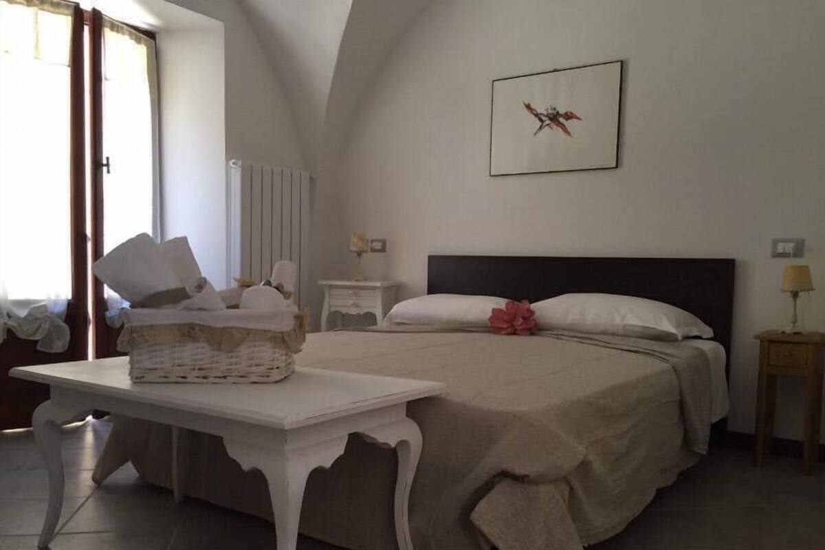 A bedroom with a bed, bedside tables, a nightstand, a window with curtains, and a framed picture on the wall.