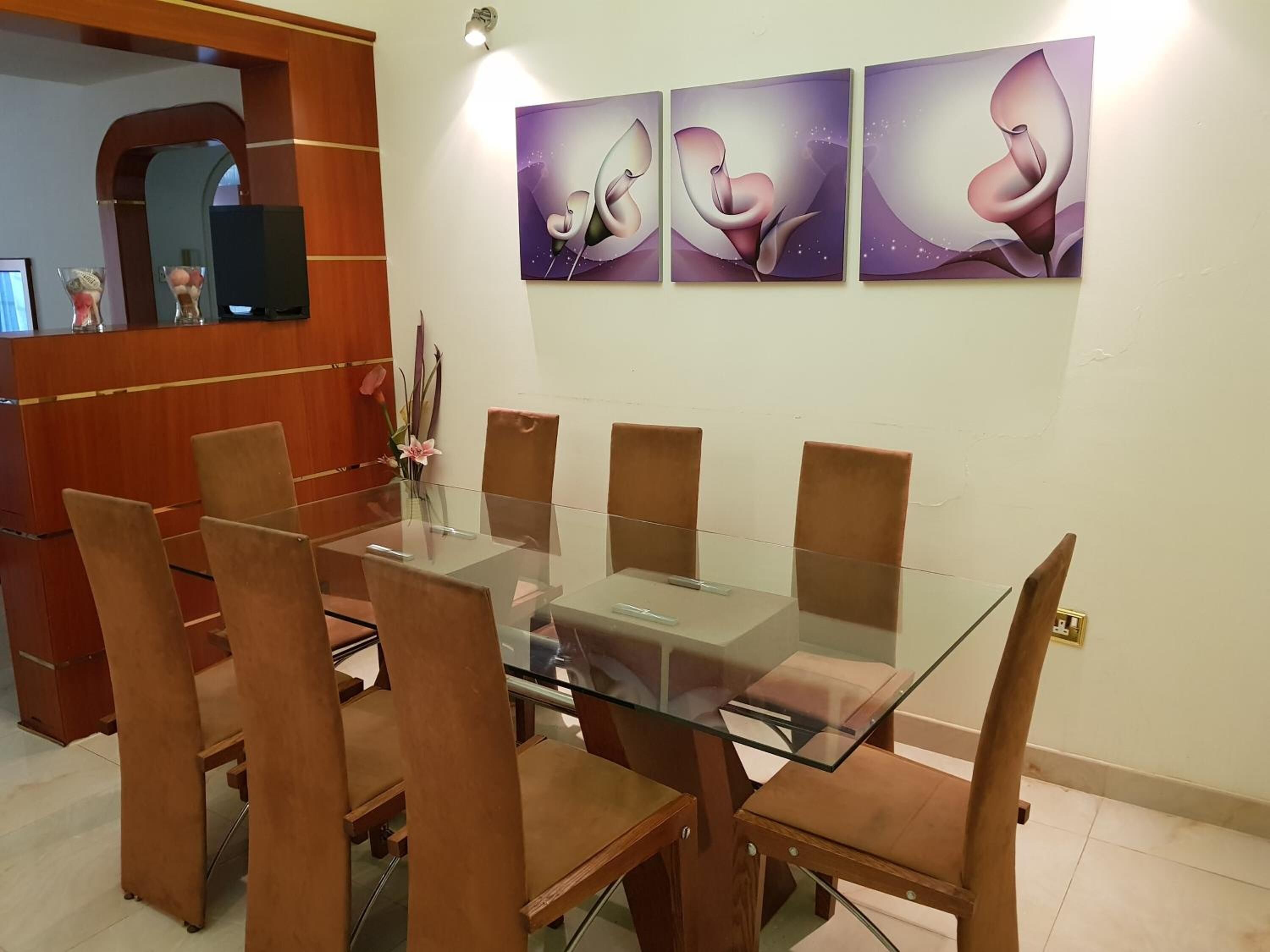 A dining area with a glass-top table surrounded by six brown chairs. There are three framed abstract paintings on the wall above the table.