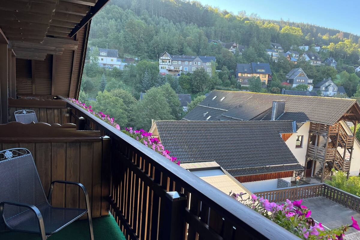 Un balcon avec des balustrades en bois, une chaise et une vue sur des maisons et des arbres.