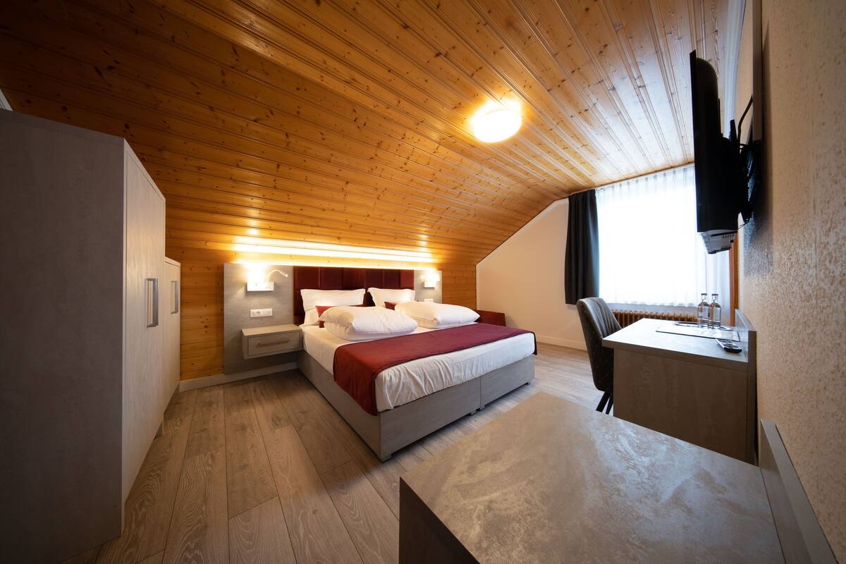 Une chambre avec un plafond en bois, un lit avec une literie blanche, un bureau avec une chaise et une fenêtre avec des rideaux.