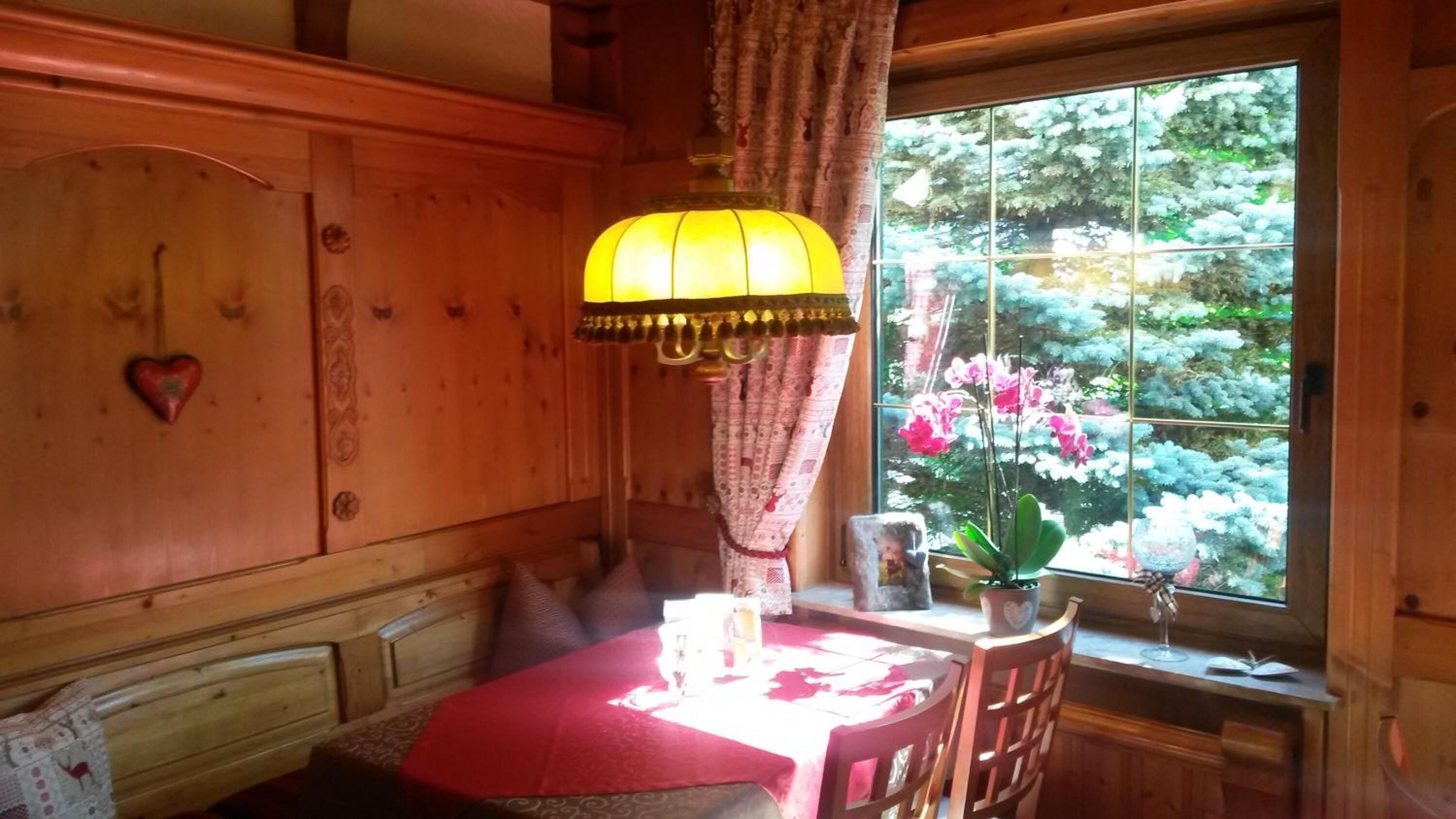 A wooden room with a dining table, a hanging lamp, a window with a view of flowers, and a heart-shaped decoration on the wall.